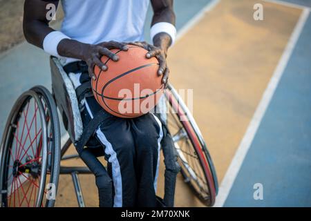 African American basketball player on wheelchair waiting to play on open air ground, concept of accessibility to sports for disabled athlete, social i Stock Photo
