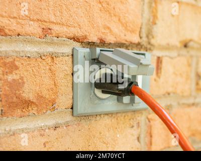 Power cord plugged into outdoor electrical wall socket. Dual covered electricity power outlets on brick wall. Orange extension cord plug Stock Photo