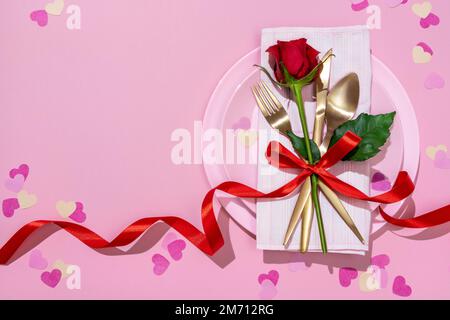Serving table with golden fork, knife, spoon and red rose tied with red ribbon on pink dish. Valentine's Day or festive romantic dinner. Stock Photo