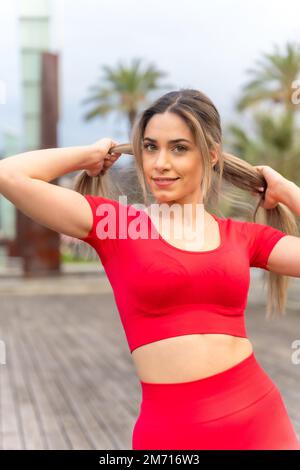 Young fit woman in red clothes doing sports in the city in spring, fitness, putting on ponytail Stock Photo
