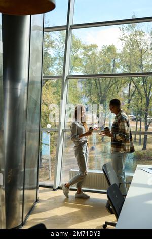 Two colleagues holding coffee while talking near big window Stock Photo