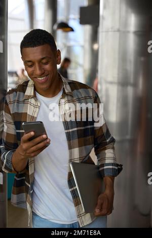 Smiling guy reading the message on phone while holding laptop Stock Photo