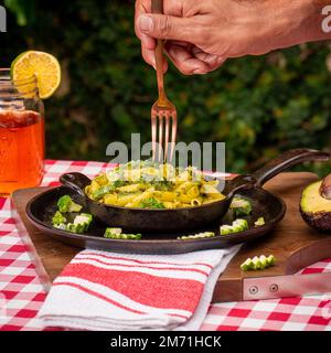 A man trying to eat the macaroons in a black bowl seasoned with greens and other ingredients on a wooden board next to a glass of juice in the yard Stock Photo