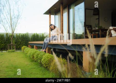 Portrait of pregnant Happy Asian woman sitting on terrace at home and using smart phone for listening music in headphones. Stock Photo