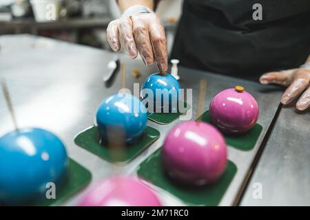 Three blue and three pink cakes on green square plates being decorated by a expert wearing transparent gloves and black apron. Blurred foreground. High quality photo Stock Photo