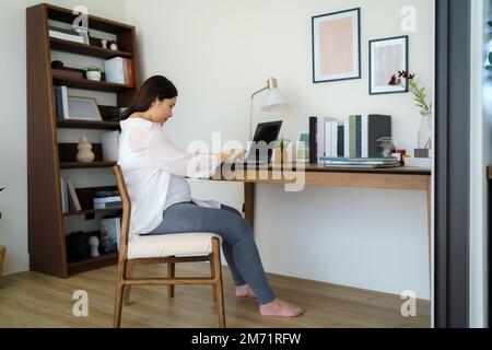 Asian pregnant woman  using a laptop while working on maternity leave at her living room table at home. Stock Photo