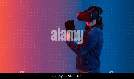 Smiling beautiful young latin woman wearing virtual reality goggles standing facing the camera, holding her plastic game gun illuminated with blue and Stock Photo