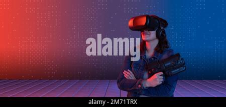 Smiling beautiful young latin woman wearing virtual reality glasses standing looking up, arms crossed, holding plastic game gun in her hand illuminate Stock Photo