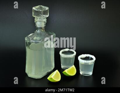 Two shots of tequila with salt around the edges and a bottle of alcohol on a black background, next to pieces of ripe lime. Close-up. Stock Photo