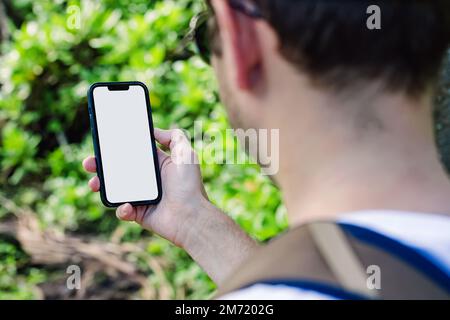 Man holding a smart phone with a blank white screen on the background of blurred lush greenery. Phone mock-up Stock Photo
