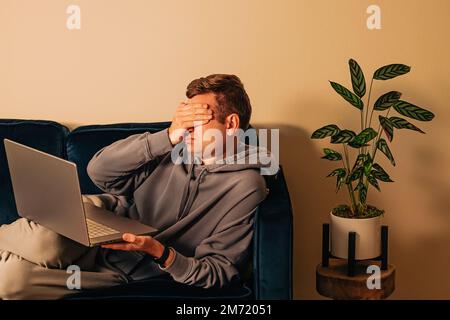 Frustrated man lying on the couch with laptop. Covering face with his hand, sadness, frustration, depression Stock Photo