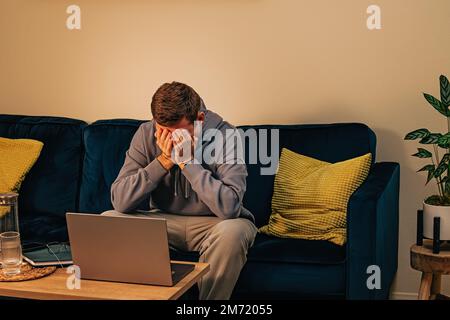 Frustrated man sitting on the sofa in front of his laptop. Covering face with his hands, sadness, frustration, depression Stock Photo
