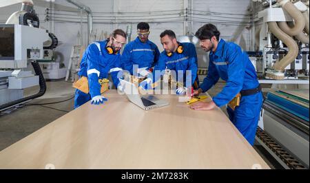 Group of multiethnic technicians in blue mechanic jumpsuits meeting,  using laptop computers on large wooden table. Production line atmosphere in furn Stock Photo