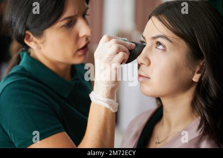 Close up of female beauty specialist using blending sponge while applying foundation under woman eye. Professional makeup artist doing makeup for client in visage studio. Stock Photo
