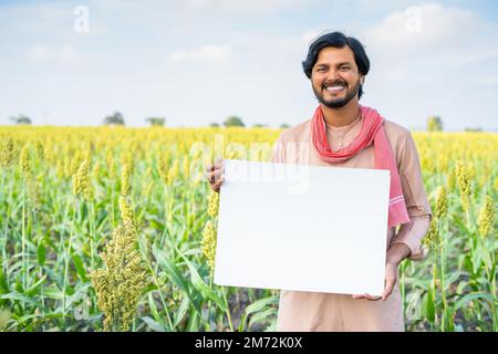 happy smiling young indian farmer holding empty sign board at agricultural farmland - concept of advertising, promotion and occupation Stock Photo