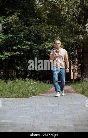 Stylish, confident mature gray haired man in sunglasses with jacket on shoulder walk street in park. Weekend activity Stock Photo