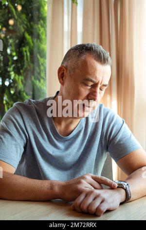 Busy grizzled mature man in gray t-shirt, look at time on wristwatch, sit at table near window, waiting for candidate for interview job Stock Photo