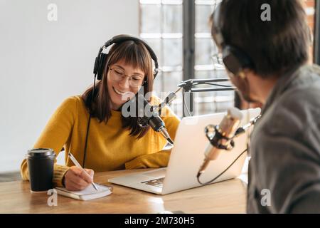 Female podcaster in headphones interviewing man guest using microphone and laptop, writing in notebook, smiling, recording and broadcasting audio podcast in studio. Content creator, blogging Stock Photo