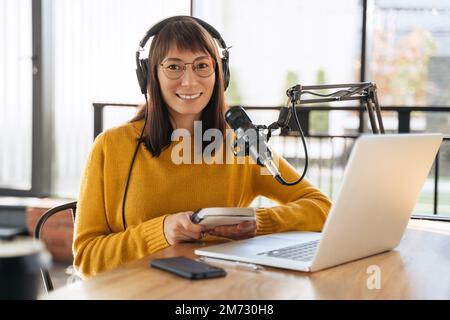 Portrait of cheerful woman host streaming audio podcast using microphone and laptop in broadcast studio, holding notepad, looking at camera and smiling. Young female podcaster running her radio show Stock Photo