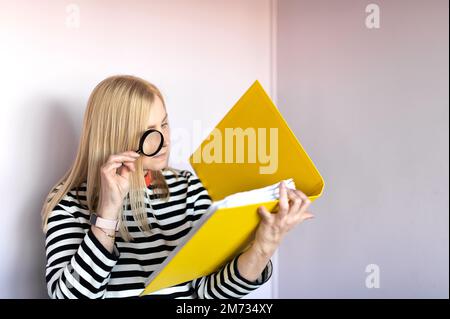 Middle-aged woman  standing isolated over light background and reading document with magnifying glass Stock Photo