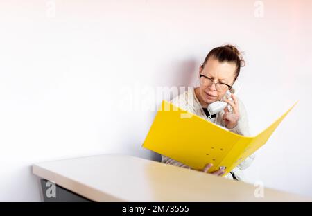 A tired-looking middle-aged woman talks on phone and hold a folder of documents at her workplace Stock Photo