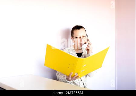 Smiling middle-aged woman talks on phone and hold a folder of documents at her workplace Stock Photo
