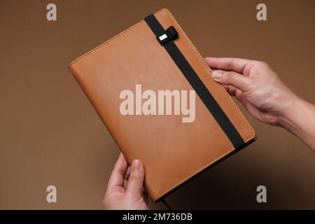 Hands hold large tan leather journal with black strap against brown surface. Planner for new year. Classy executive organizer. Horizontal background. Stock Photo