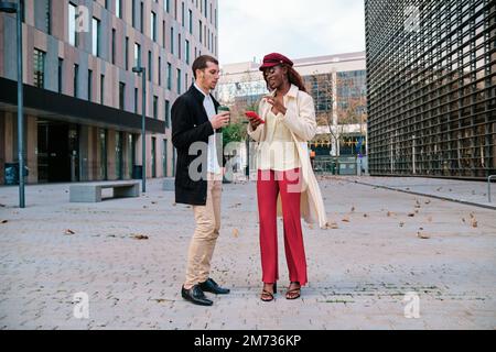 Two business people drinking coffee and using mobile phone outdoors in the financial district. Stock Photo