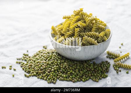 Mung bean fusilli pasta on a gray textile background. Bowl with raw pasta and green mung bean. Gluten free pasta. Stock Photo