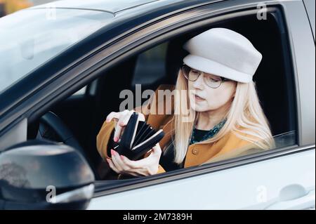 Middle-aged woman sits behind the wheel of car and looks for money in wallet to pay for parking Stock Photo