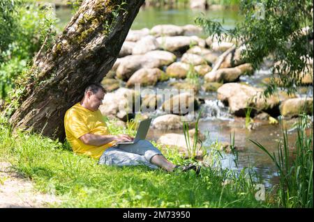 Middle aged freelancer using a laptop computer while working in beautiful nature under the tree on the bank of the river Stock Photo