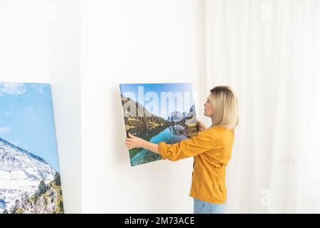 Canvas print with gallery wrap interior decor. Woman hangs landscape photography on white wall. Hands holding photo canvas print Stock Photo