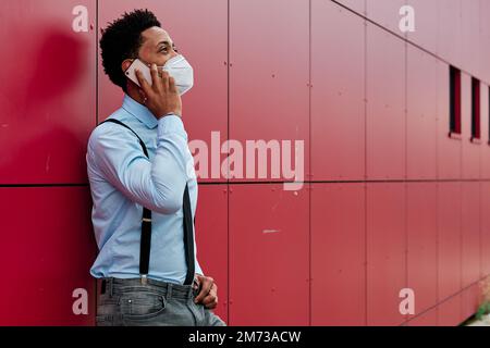Executive man with face mask talking on the phone while leaning on a wall outdoors. Stock Photo