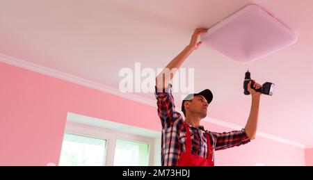 A young man stands on a stepladder and installs a large chandelier on the ceiling Stock Photo