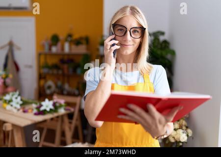 Young blonde woman florist talking on smartphone reading book at flower shop Stock Photo