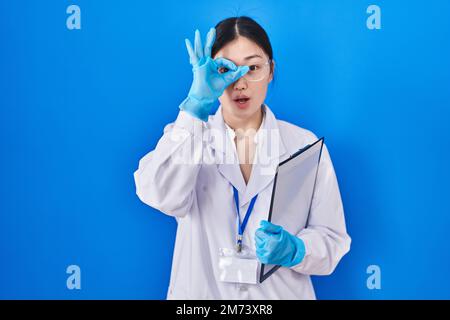 Chinese young woman working at scientist laboratory doing ok gesture shocked with surprised face, eye looking through fingers. unbelieving expression. Stock Photo