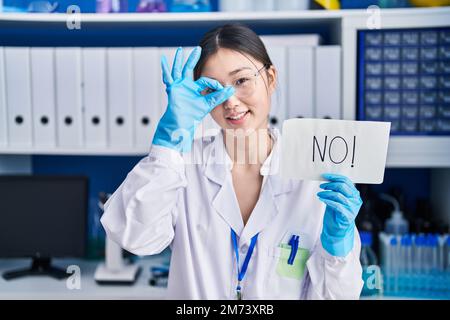 Chinese young woman working at scientist laboratory holding no banner smiling happy doing ok sign with hand on eye looking through fingers Stock Photo