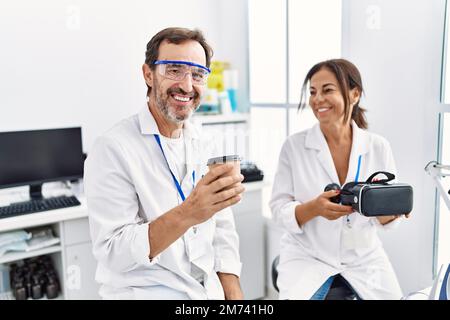 Middle age man and woman partners wearing scientist uniform drinking coffee and holding vr goggles at laboratory Stock Photo