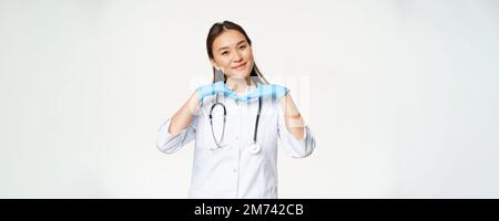 Image of smiling asian doctor, dermatologist shows healthy skin and posing cute at camera, standing in medical uniform over white background Stock Photo