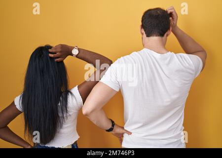 Interracial couple standing over yellow background backwards thinking about doubt with hand on head Stock Photo