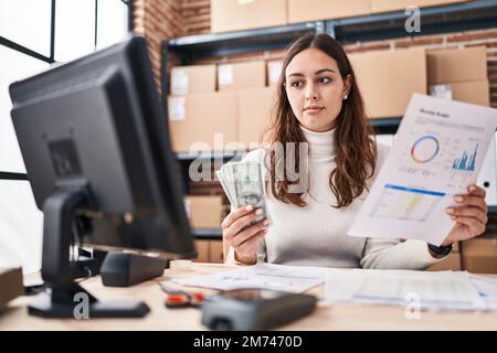 Young beautiful hispanic woman ecommerce business worker holding dollars reading document at office Stock Photo