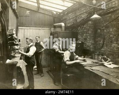 England. Circa. 1915. An interesting antique photograph of a group of craftsmen in a factory workshop setting. An older gentleman and three younger men, possibly apprentices, are carrying out their tasks. Early electric light fittings are mounted on the wall and suspended from the ceiling. Stock Photo