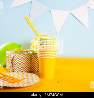 Accessories for a party or birthday on yellow background. Paper straws, wrapped gift box, cups, flags, plates. Stock Photo