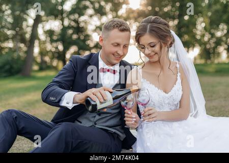 Smiling and shy groom and bride drinking and pouring bubbly champagne wine from bottle to glasses together, sit in park Stock Photo