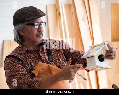 Happy Asian senior man is enjoying his hobby by diligently painting birdhouses in the workshop. Stock Photo