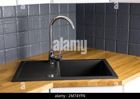 Close up view of new modern kitchen solid wood butcher block oak counter with built in black rectangular granite sink, repainted gray color tile wall. Stock Photo