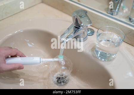 White man's hand washing the electric toothbrush in the bathroom sink. Stock Photo