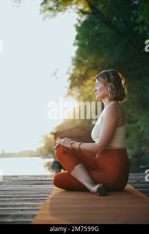 Yogi woman meditates and does breathing exercises while sitting on the dock. Stock Photo