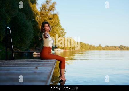 A middle-aged woman is relaxing on the dock and doing yoga breathing exercises. She put her feet in the water. Stock Photo