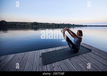 A fit yogi woman is practicing yoga on a dock at dusk. She is in the Boat yoga posture. Stock Photo